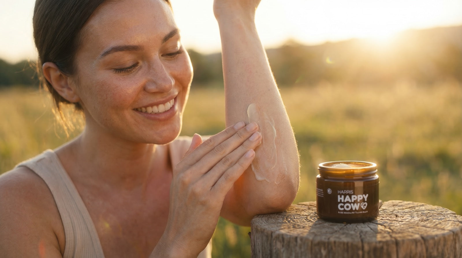 Woman applying tallow balm lotion to her arm with a jar of 'Happy Cow' cream on a wooden stump in a field.