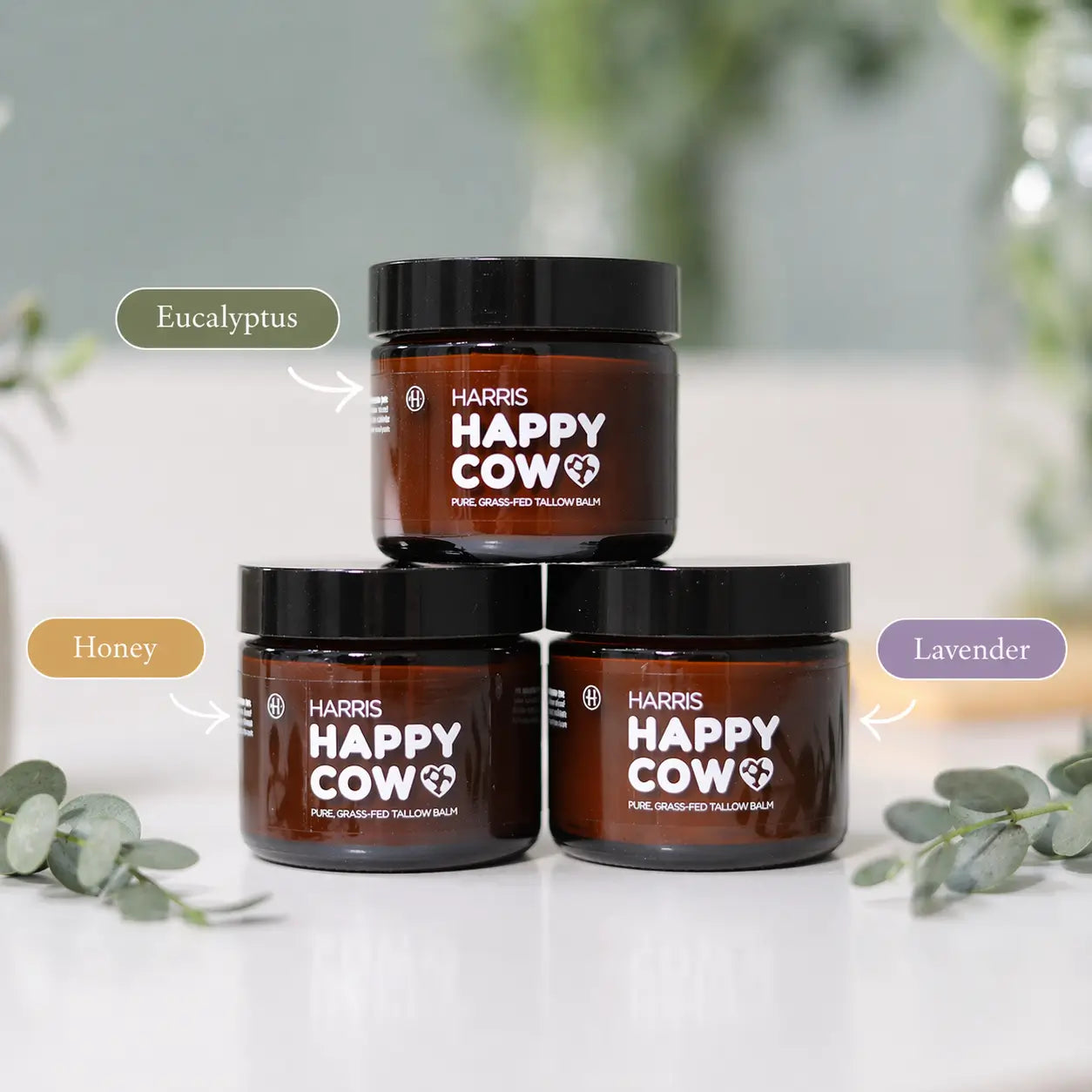 Three jars of Harris Happy Cow tallow balm on a white surface with greenery.